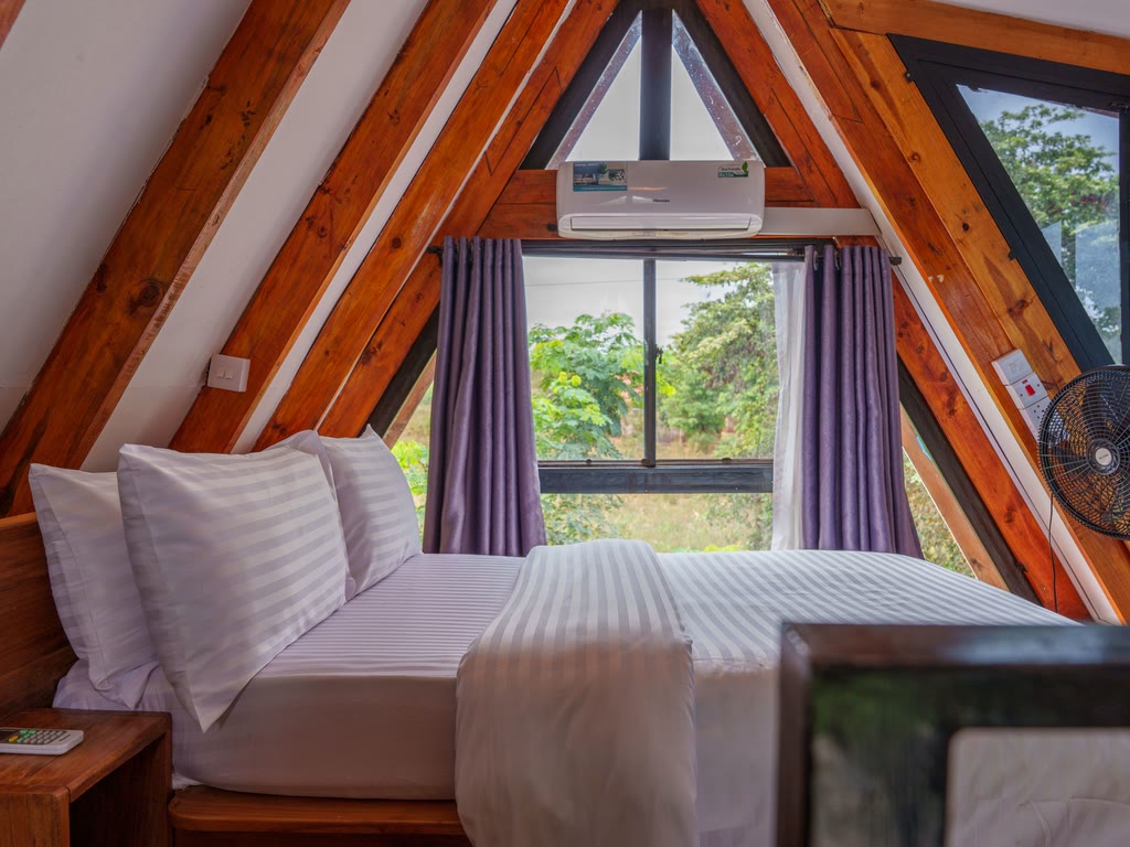 A cabin bedroom with a bed beneath timber beams, a window and an air-conditioning unit.