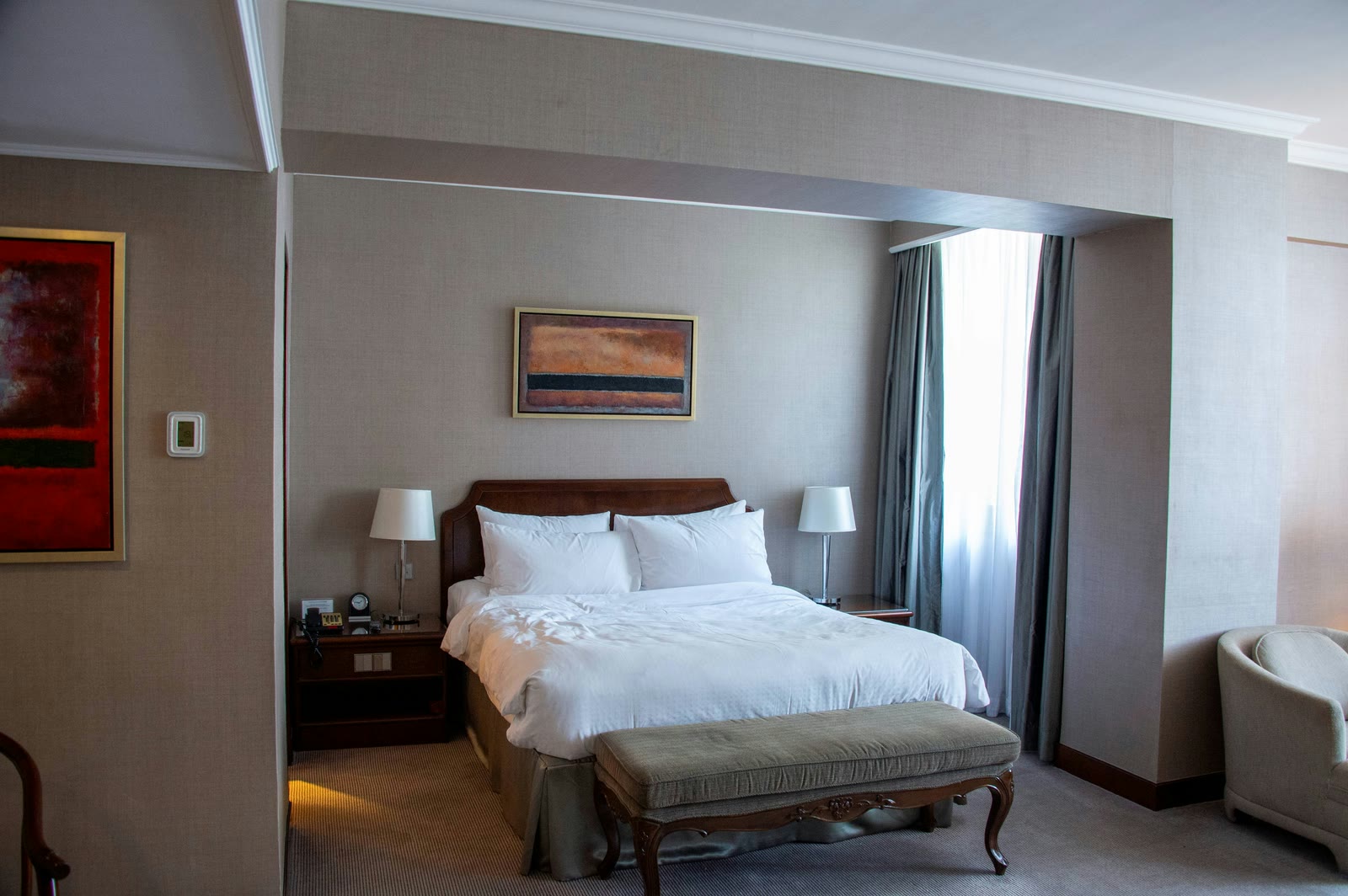 A white bed with a bench, bedside lamps and a large window.