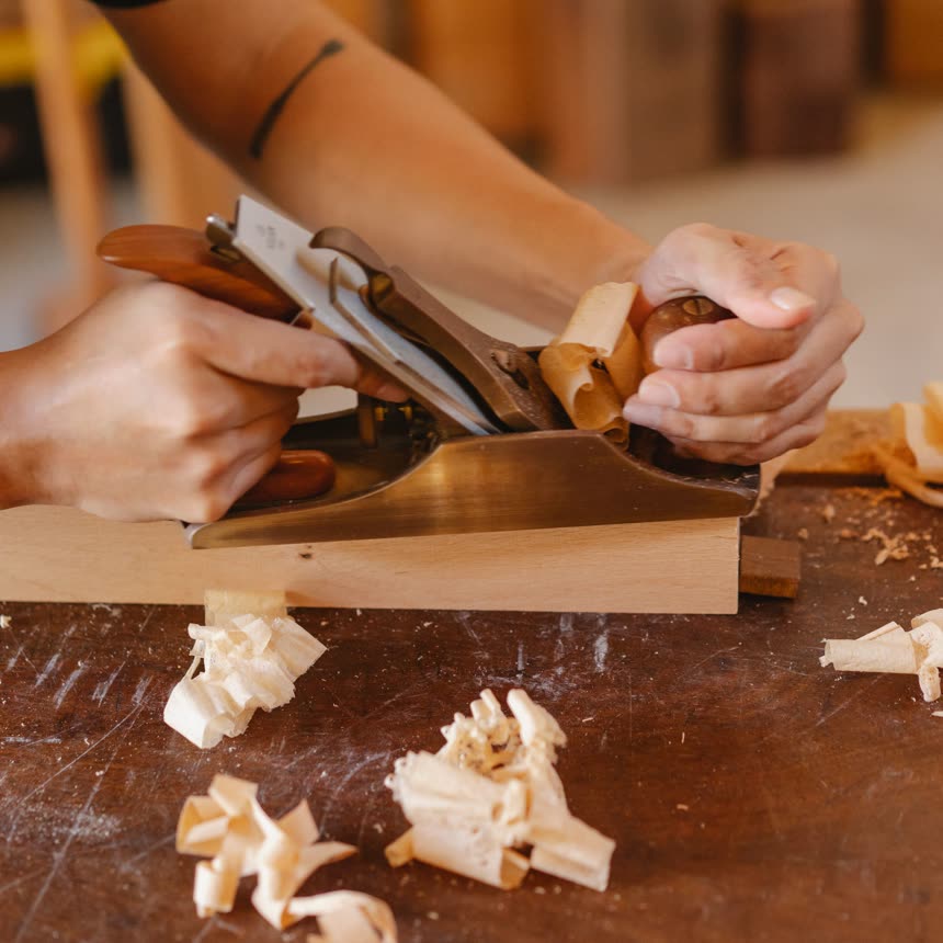 Hands pushing a hand plane, curls of wood on the bench