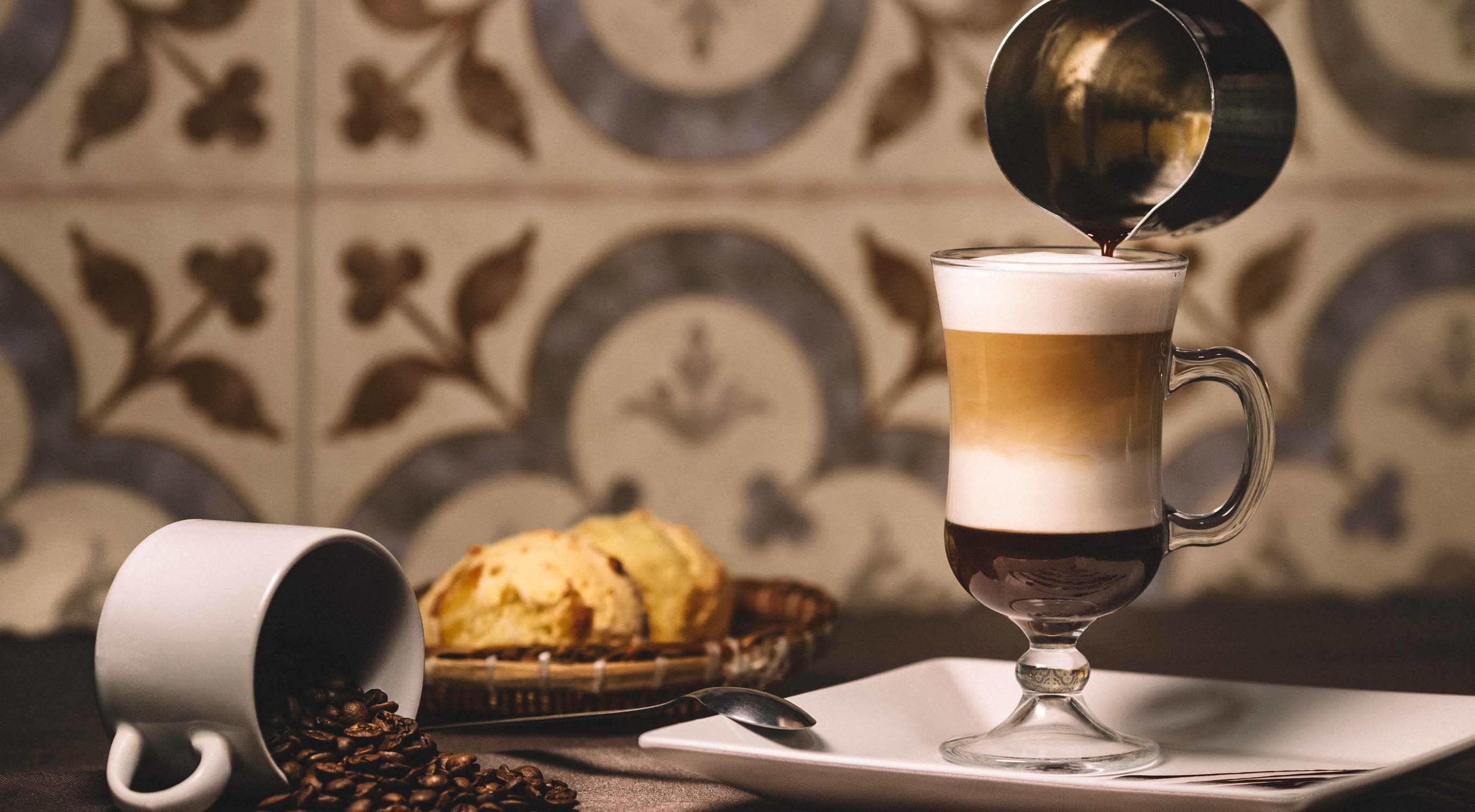 Milk poured into a layered coffee glass beside a basket of pastries, patterned cement tiles behind