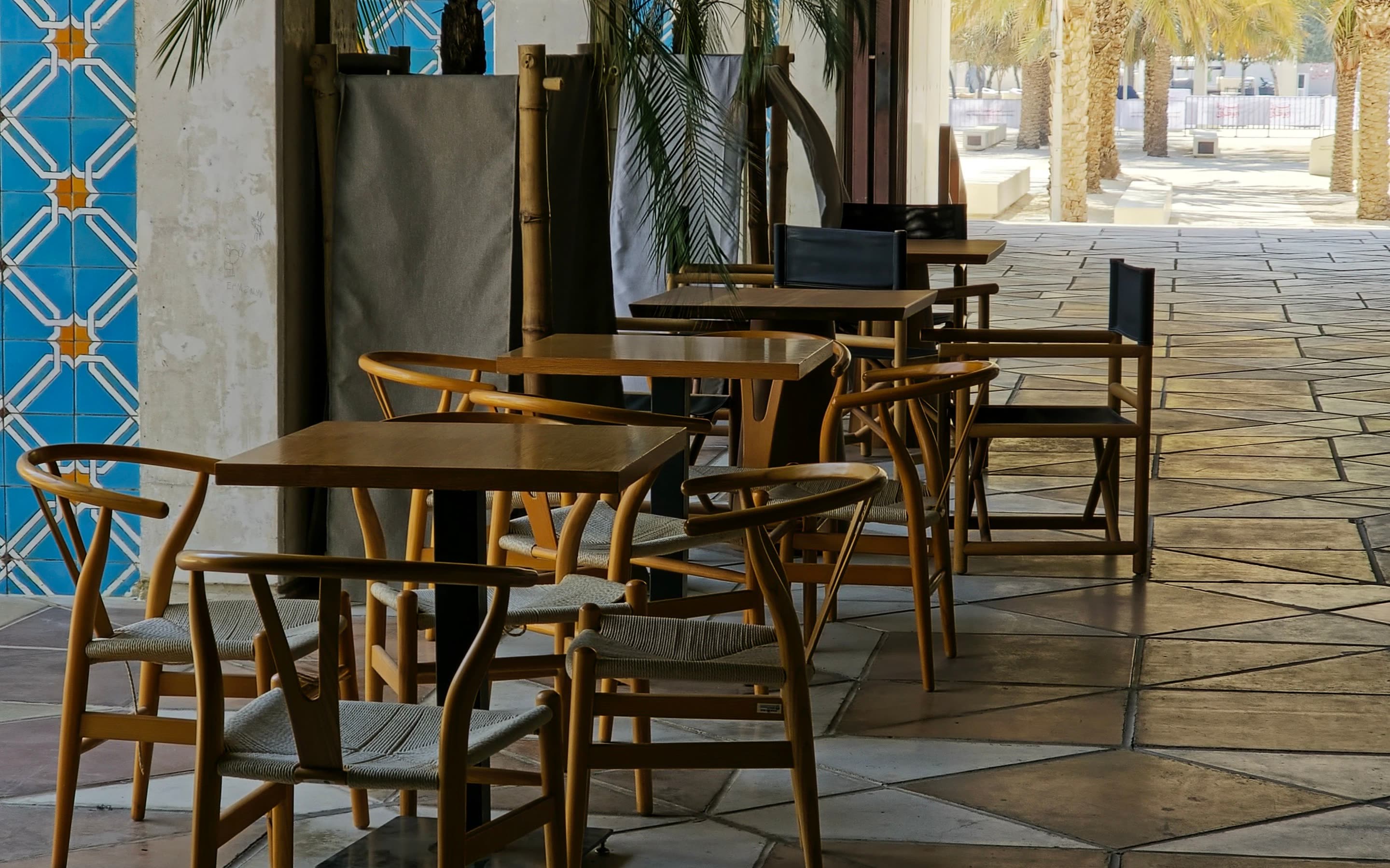 Wooden café chairs on a stone floor beside a wall of blue patterned tile and potted palms