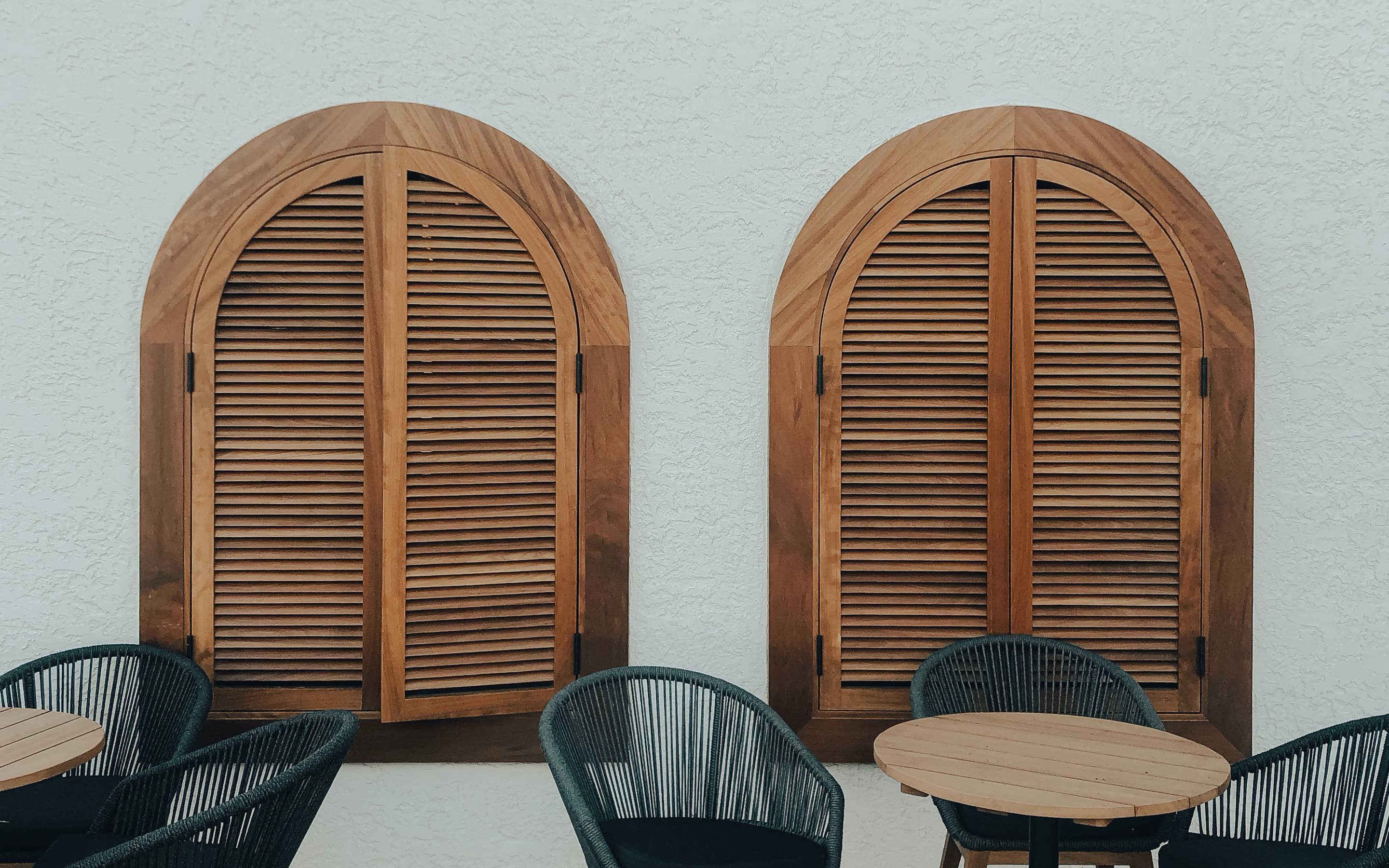 Two arched wooden shutters on a white wall above small outdoor tables
