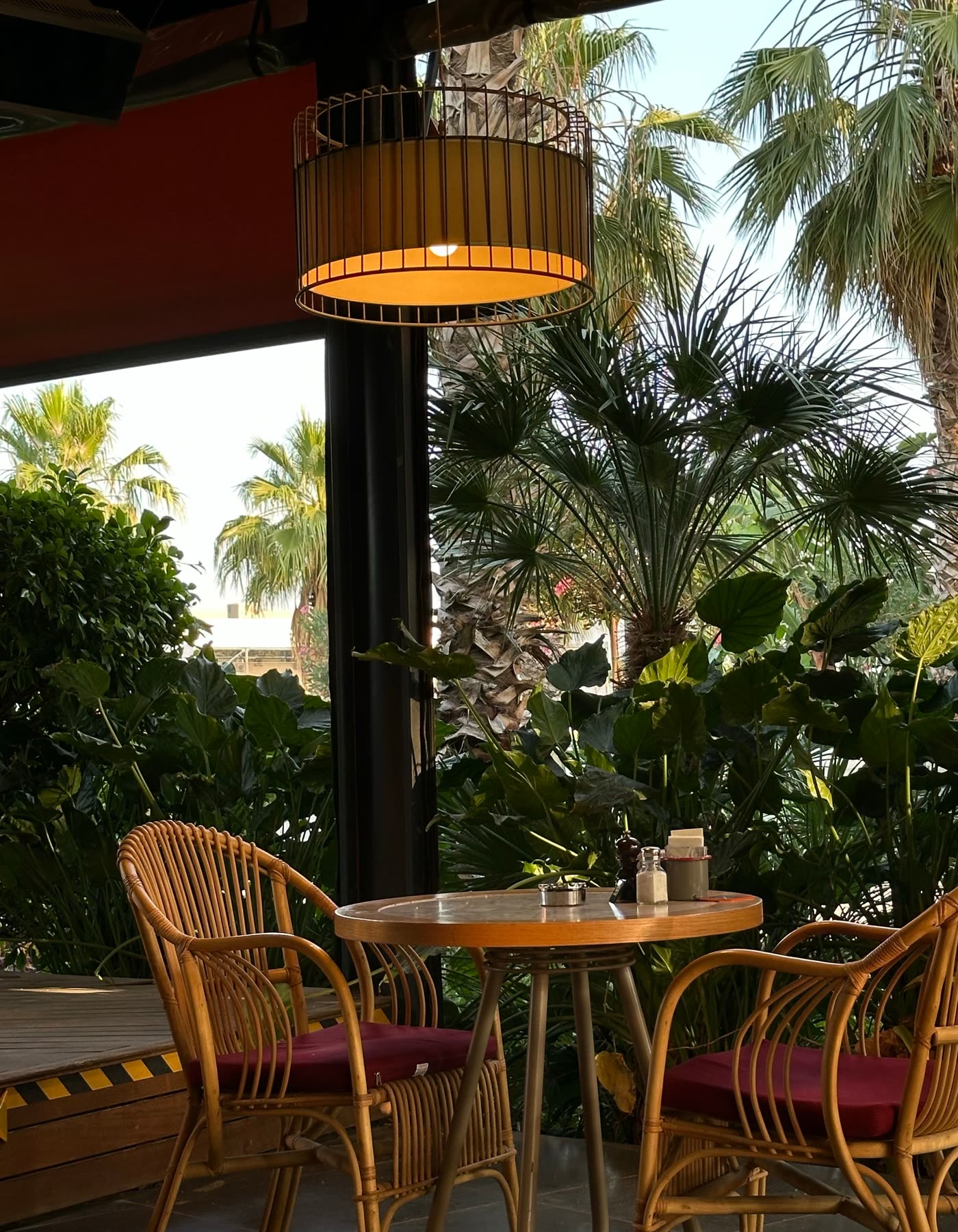Two rattan chairs and a round table under a woven lamp, palms beyond the open wall