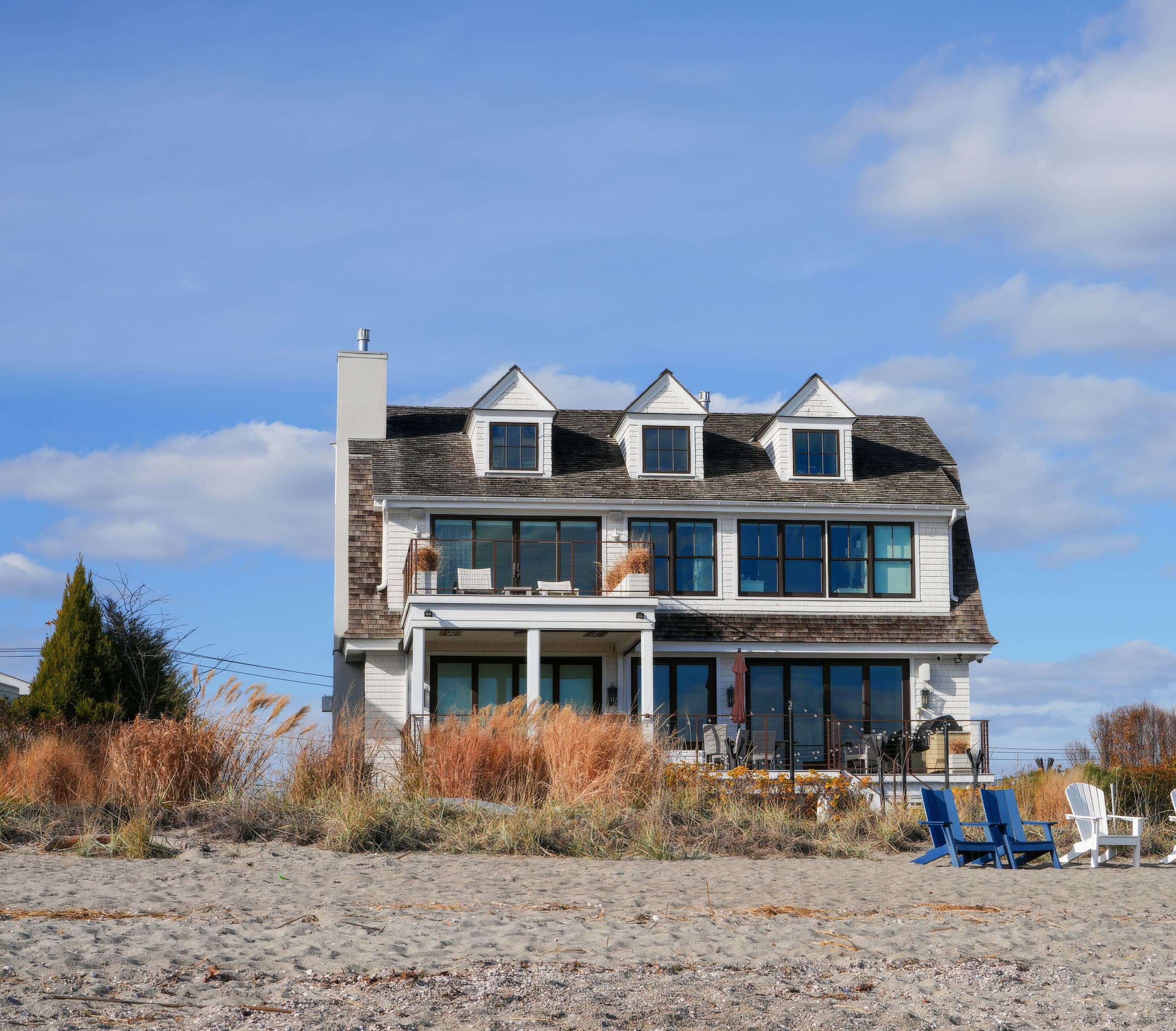 A grey shingled beach house with blue chairs on the sand in front