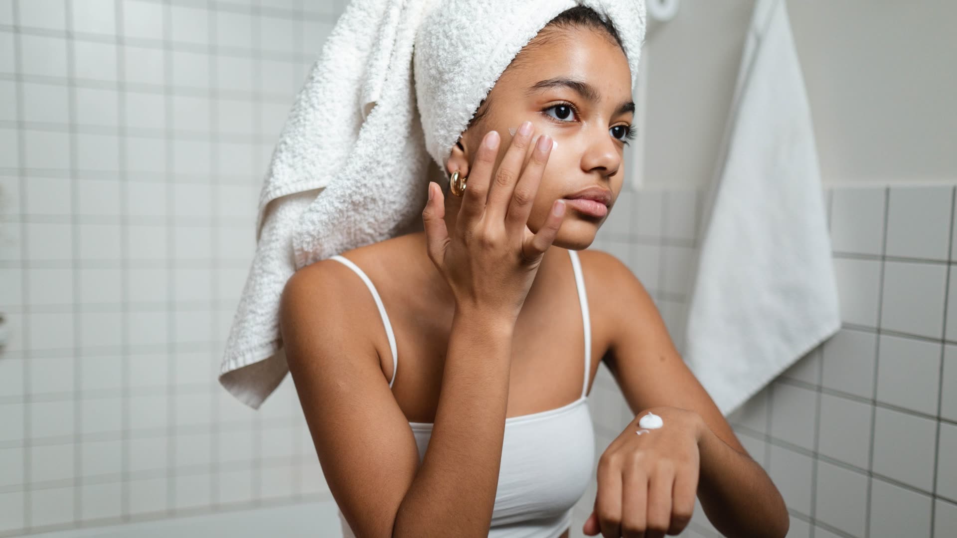 Woman with her hair in a towel applying cream in a tiled bathroom