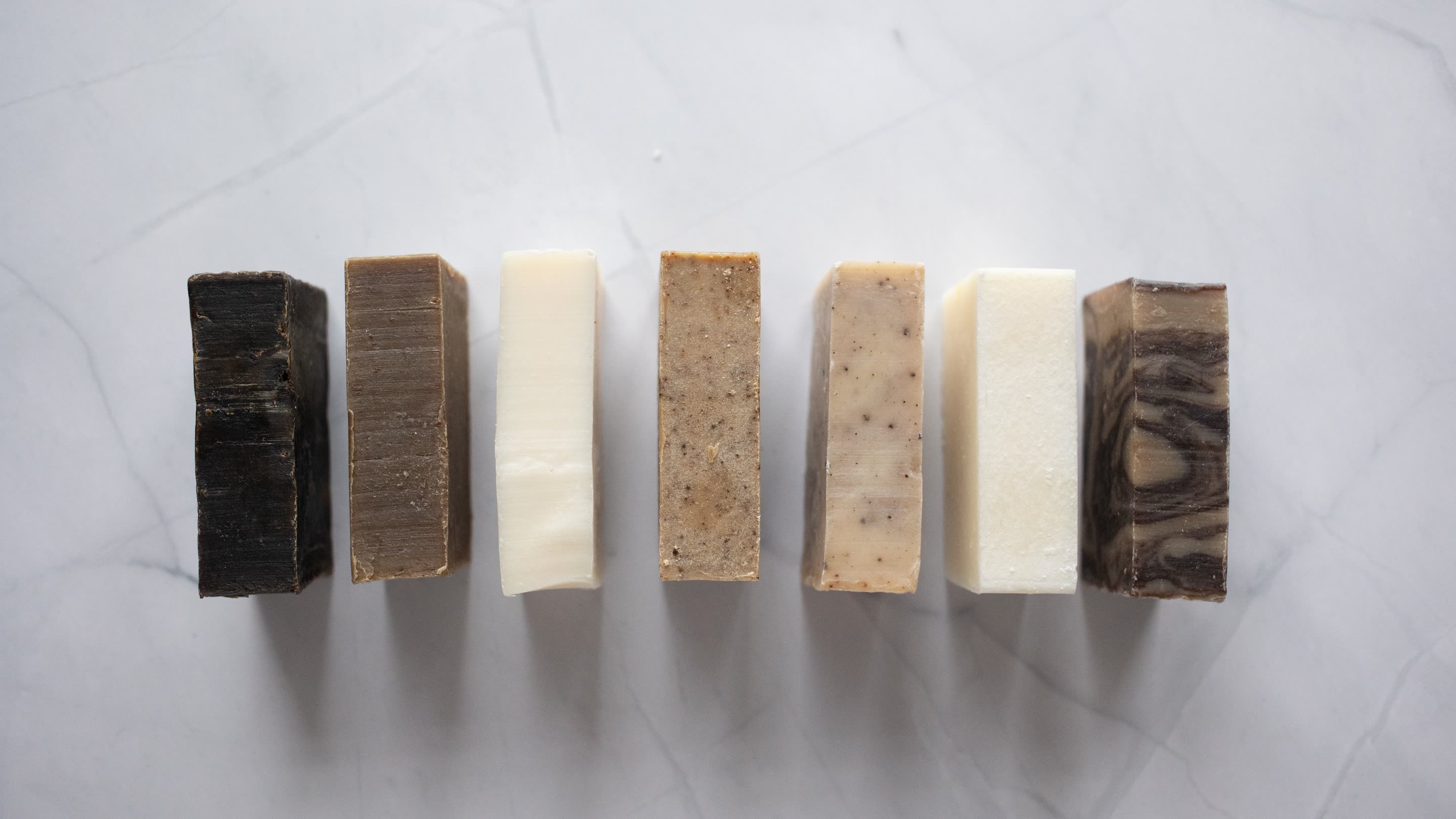 Seven handmade soap bars in a row on white marble, from dark cedar to plain shea