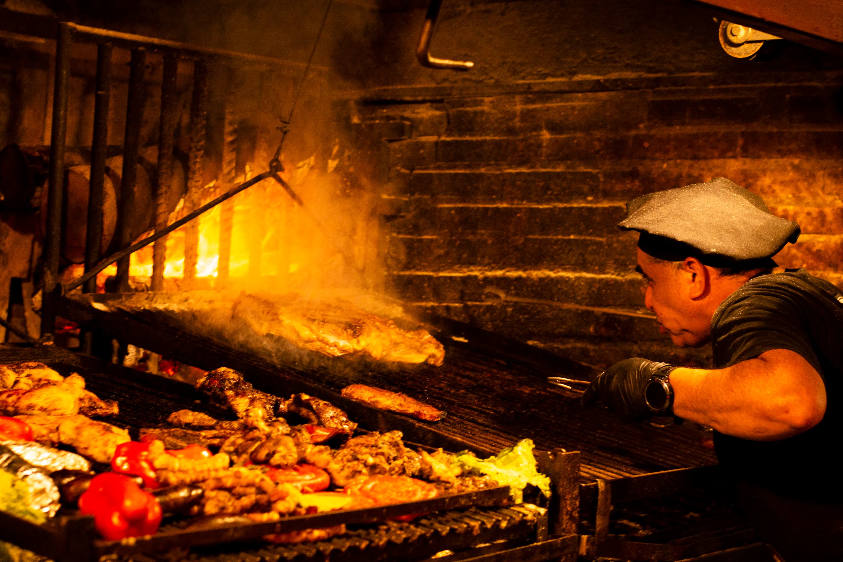 A cook tending meat and peppers over an open oak fire at a brick hearth