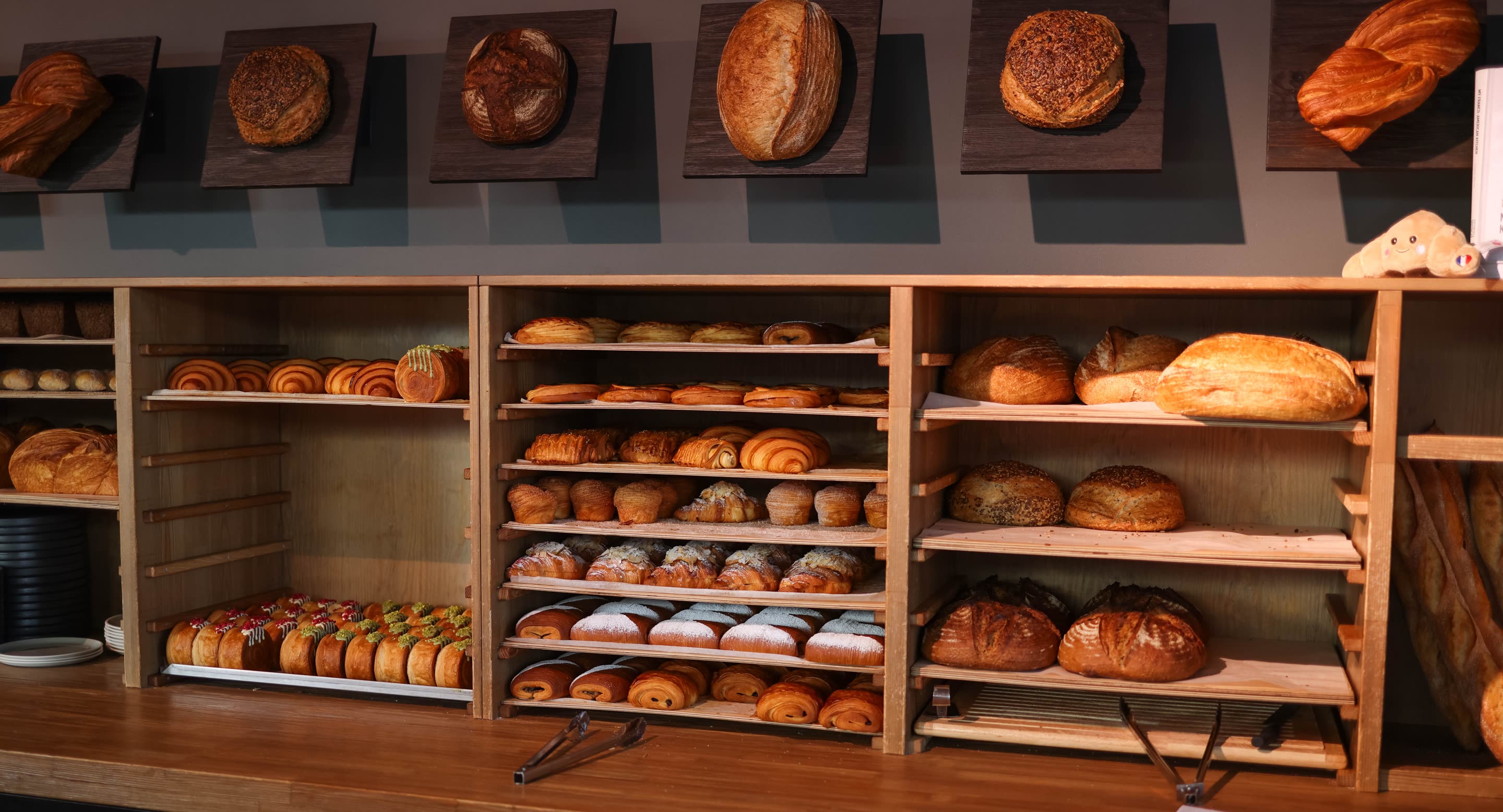 Wooden bakery shelves stacked with country loaves, rolls and morning pastry below a row of display loaves