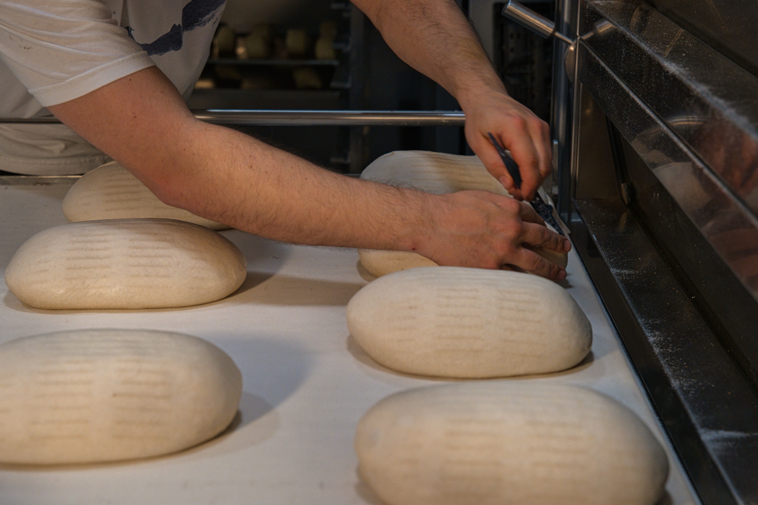 A baker scoring shaped loaves on the bench before they go into the deck oven