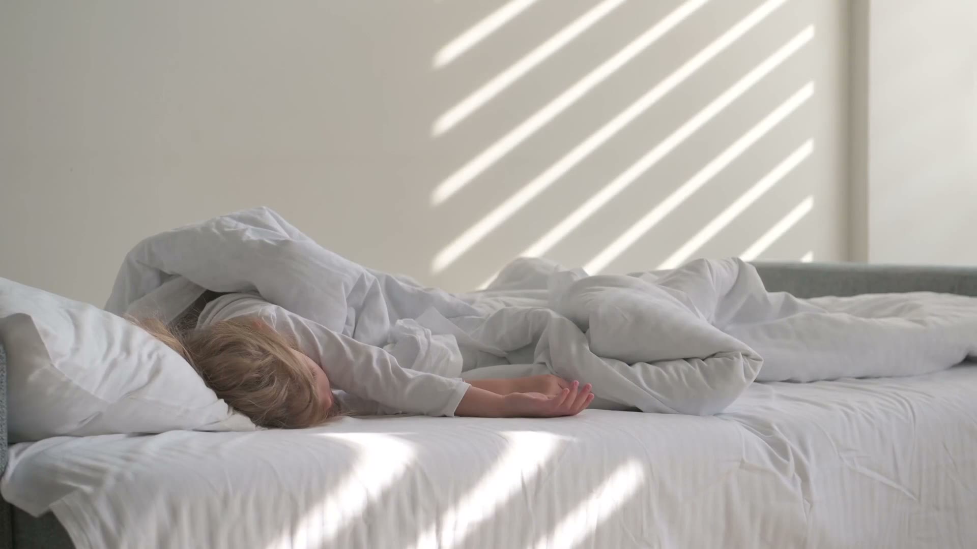 Morning light falling in stripes across a white bed where someone is still asleep