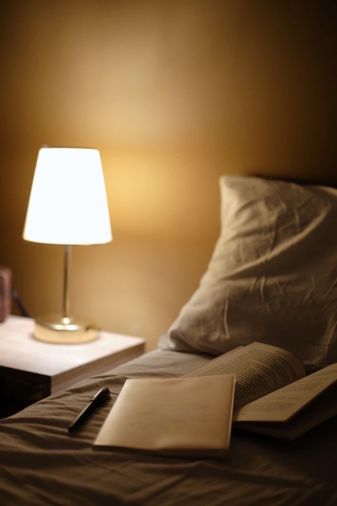 A bedside lamp lit beside a notebook on the bed