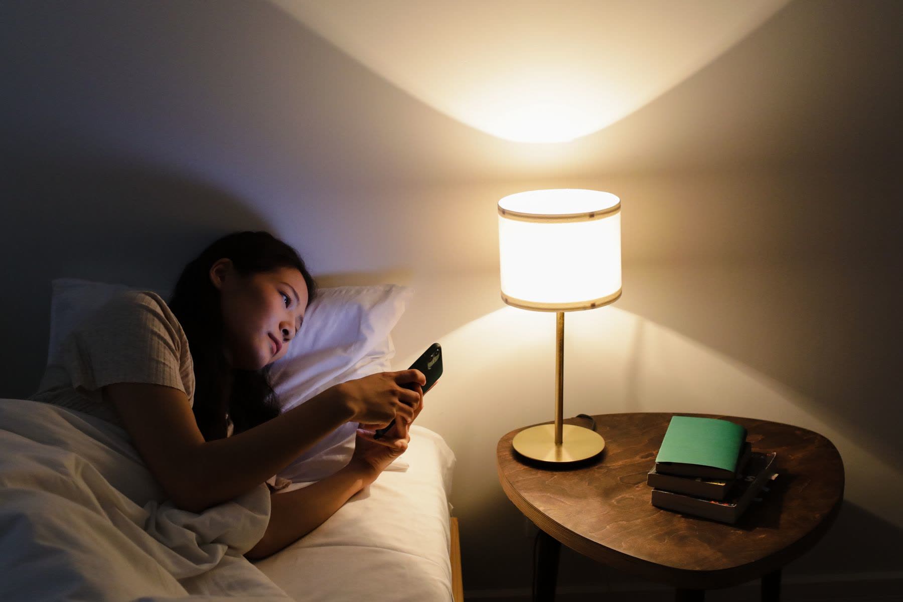 A woman in bed looking at her phone beside a lit bedside lamp
