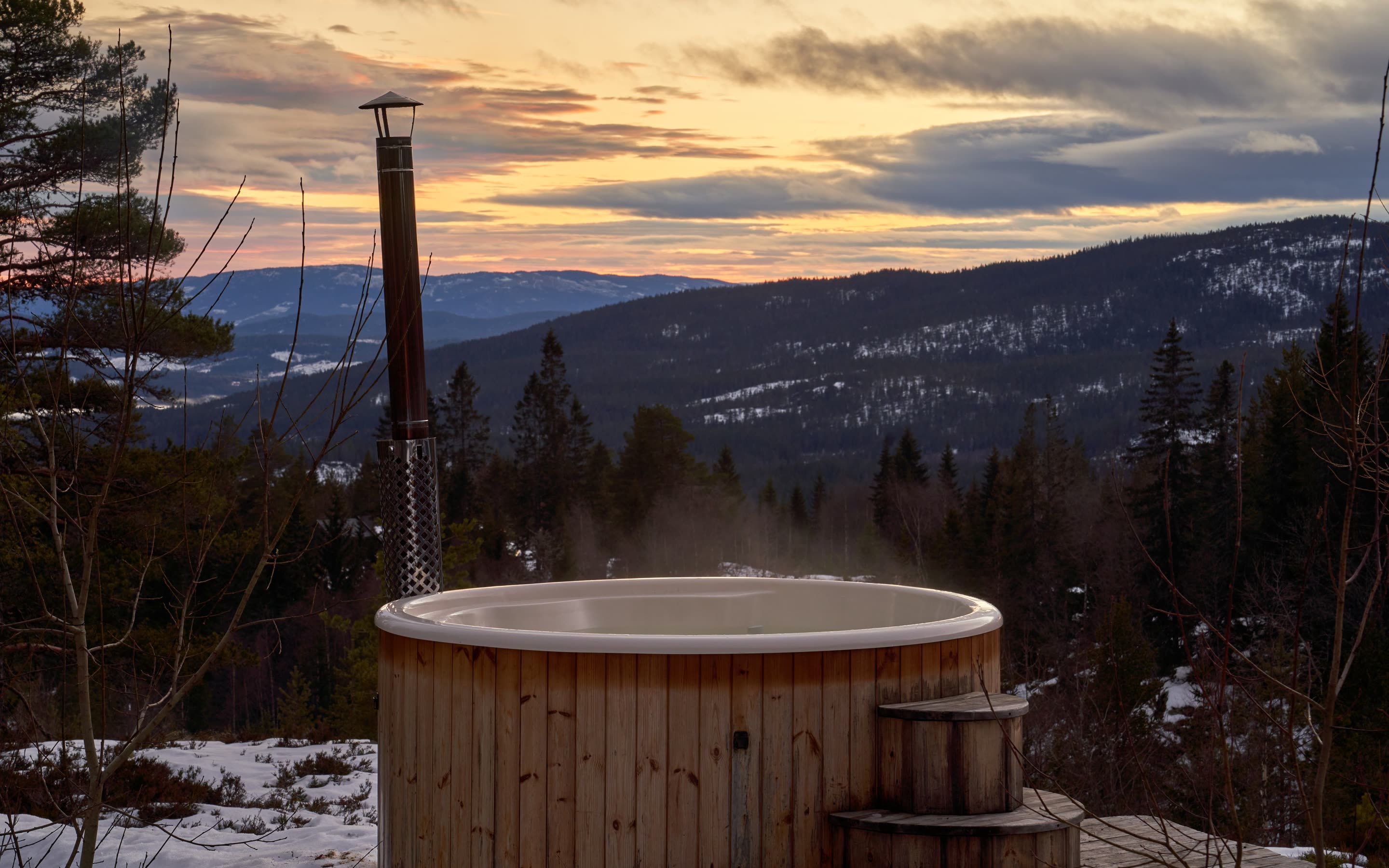 Wood-fired cedar tub steaming on a deck above forested ridges at sunset