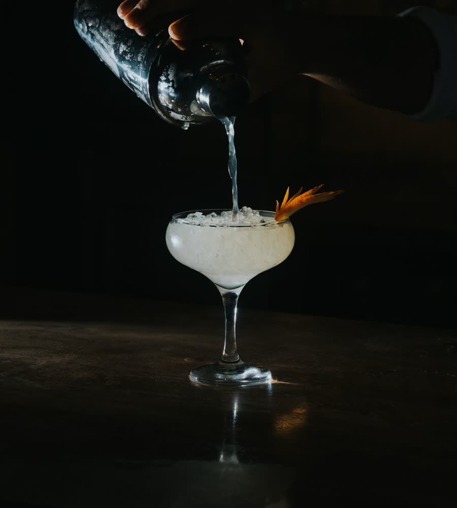 A cocktail being poured into a coupe glass at a dark bar