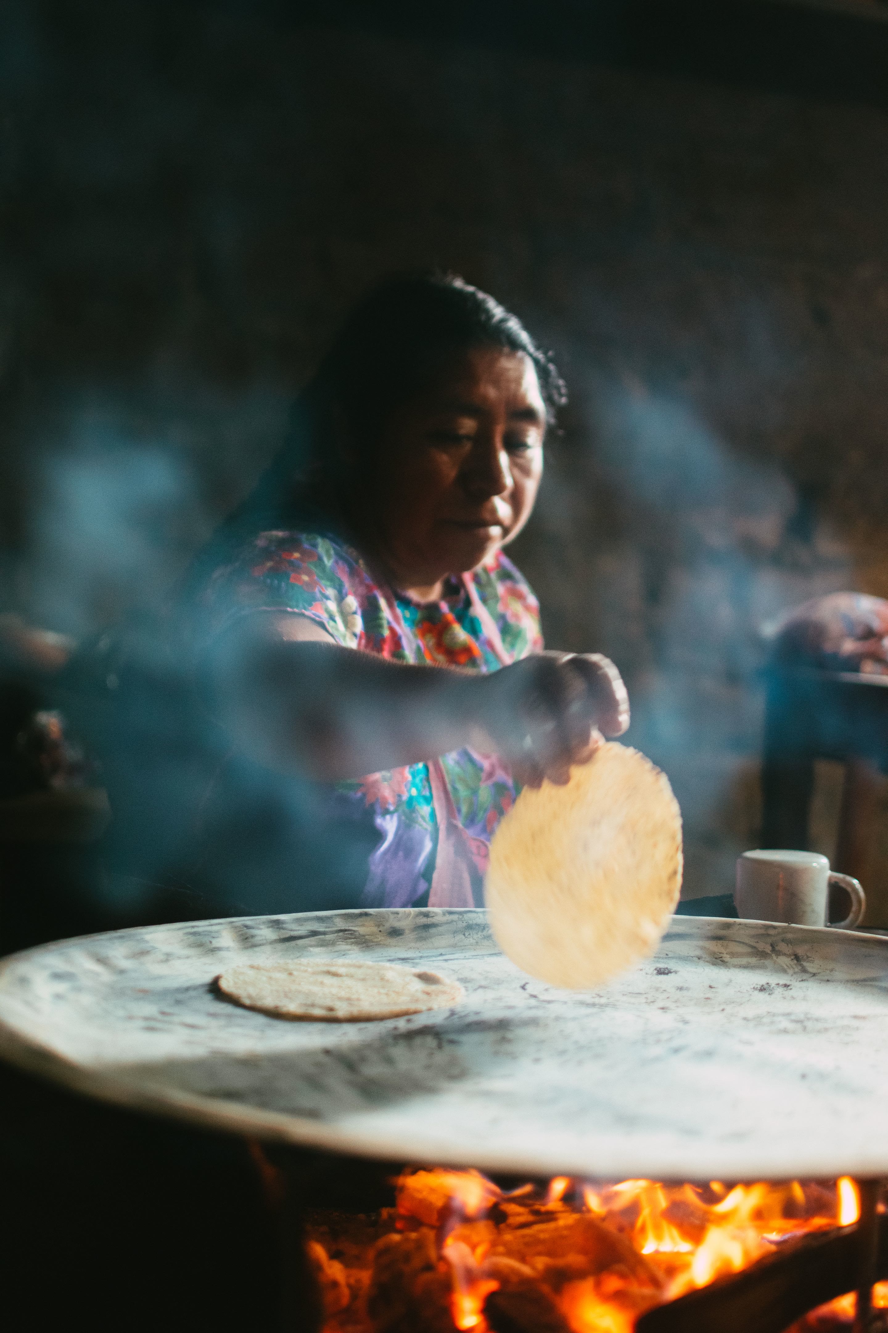 A cook lifting a fresh tortilla off a comal set over a wood fire