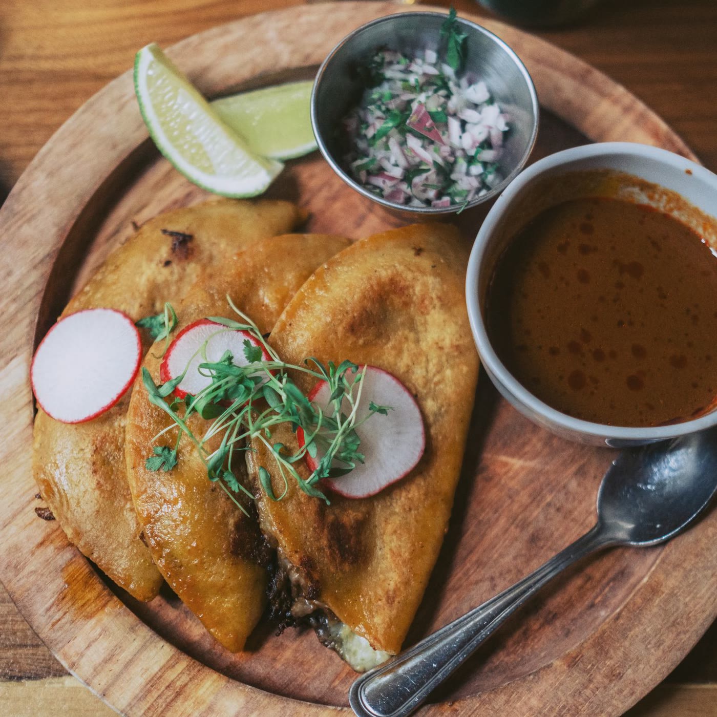 Quesabirria folded on a wooden plate with radish, lime and a cup of consomé
