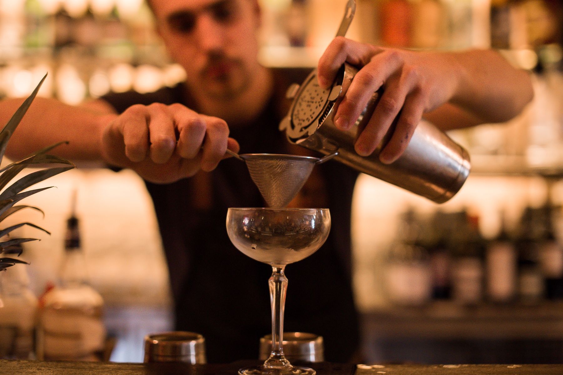 A bartender fine-straining a cocktail from a shaker into a coupe