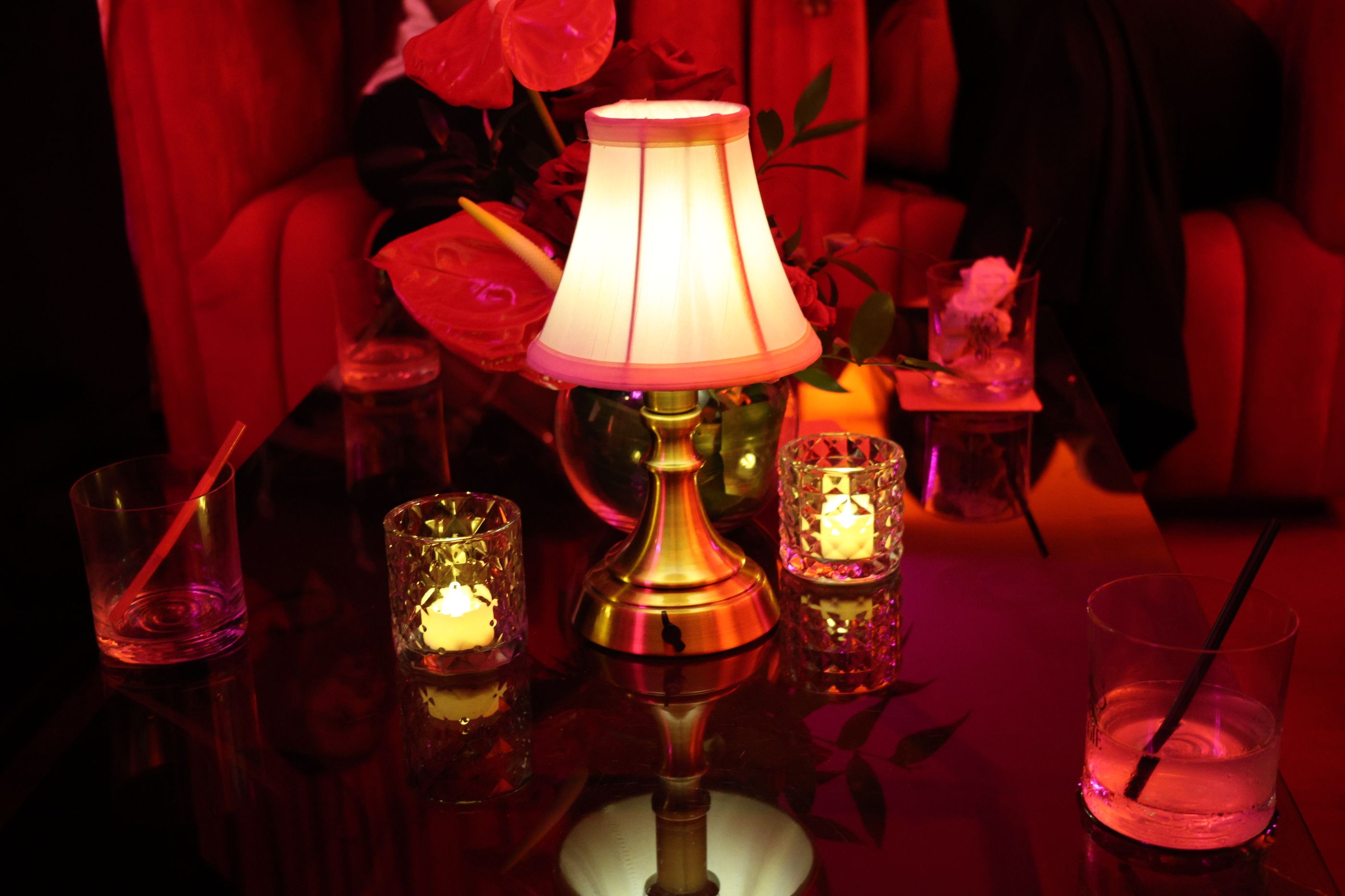 A brass table lamp, candles and cocktail glasses on a table in a red-lit lounge