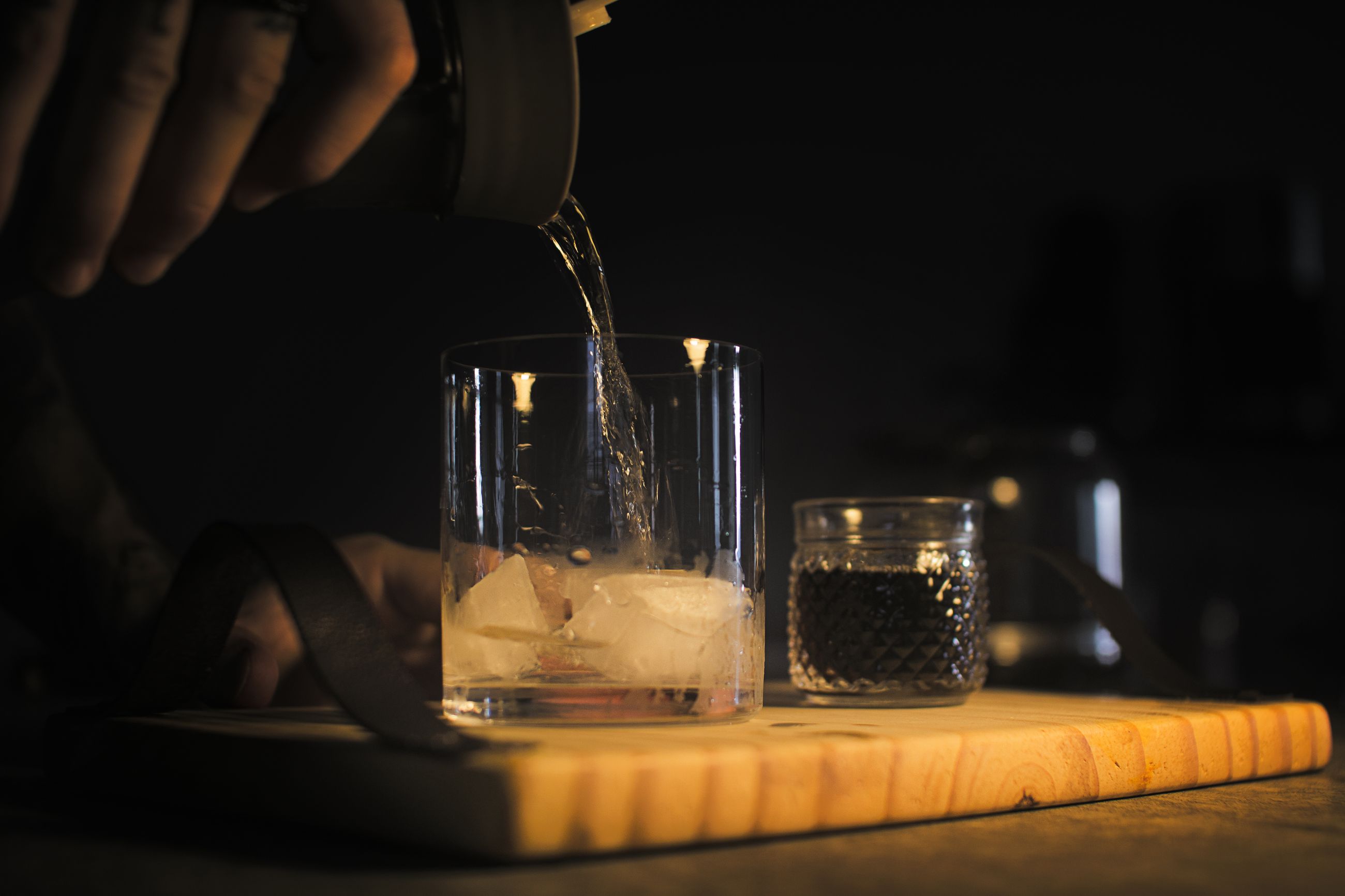 A bartender pouring whiskey over ice in a glass on a wooden board in a dark room